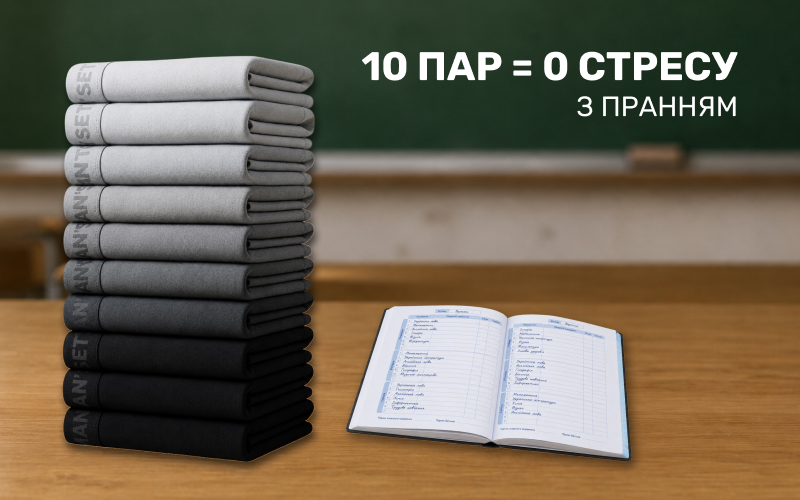 Висока стопка з десяти дитячих трусів Man's Set у градації кольорів від світло-сірого до чорного, що стоїть на дерев'яному шкільному столі поруч із розгорнутим учнівським щоденником на фоні класної дошки.