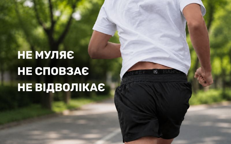 Хлопець біжить парковою дорогою, вид ззаду. Він одягнений у чорні спортивні шорти та білу футболку, з-під яких помітна еластична брендована резинка трусів Man's Set, що надійно фіксується на тілі.