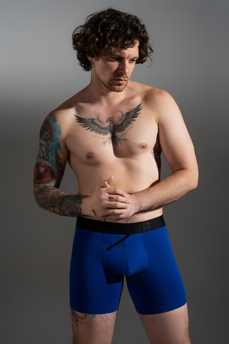 Мужчина с татуировкой феникса на груди в ярко-синих анатомических боксерах на сером фоне.